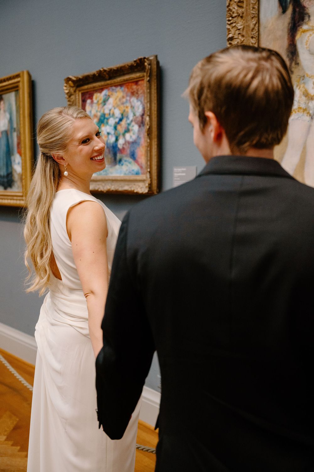 A couple interacts playfully in an art gallery with ornate gold-framed paintings on blue walls.