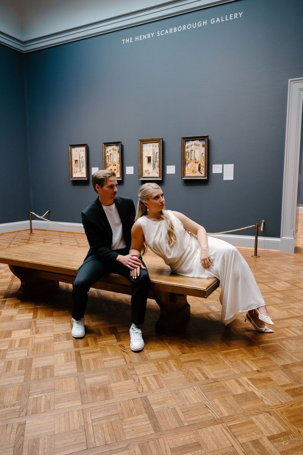 Two people sit together on a wooden bench in an art gallery with blue walls and framed artwork.