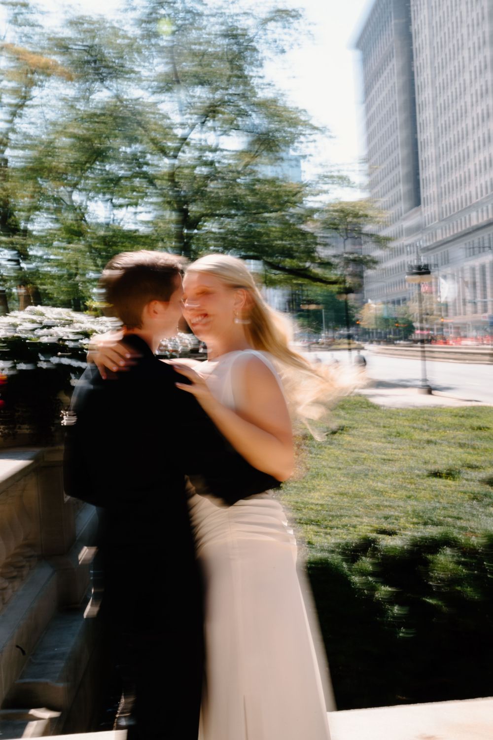 A romantic couple embraces outdoors with motion blur and urban architecture in the background.