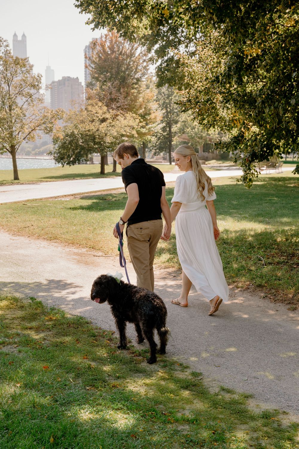 Two people walk a black dog along a tree-lined path with a city skyline in the background.