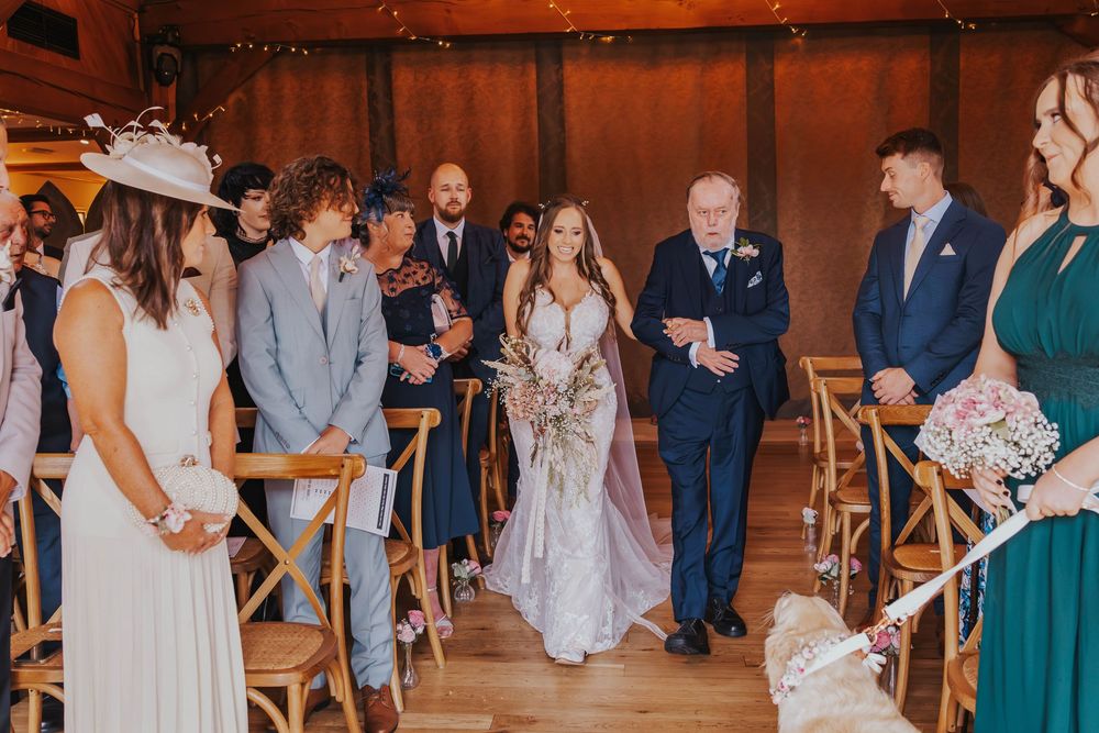 A wedding ceremony takes place in a rustic wooden venue with guests standing beside cross-back chairs in the aisle.