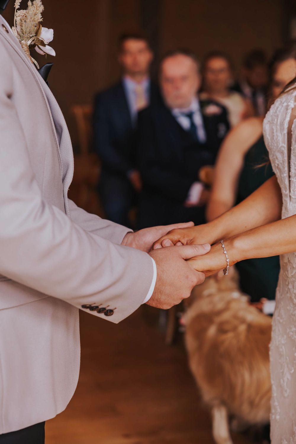 Close-up view of hands clasping during a wedding ceremony with white formal attire and guests in background.