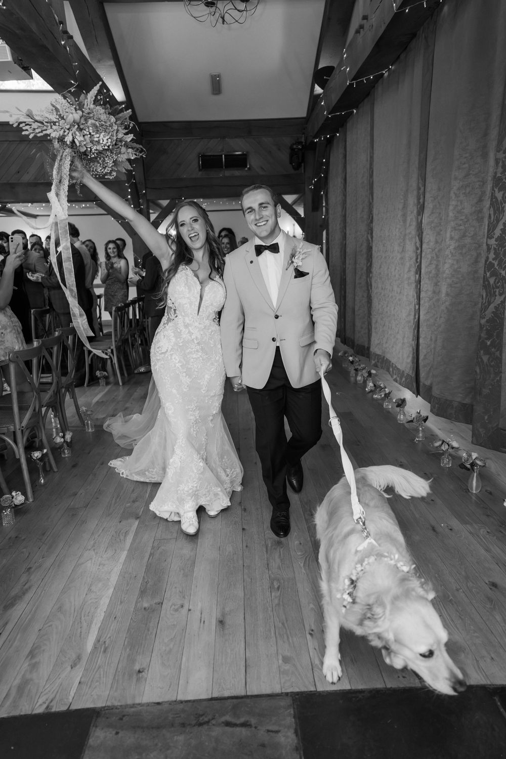 A couple in white wedding attire dances with their golden retriever on a wooden dance floor at their reception.