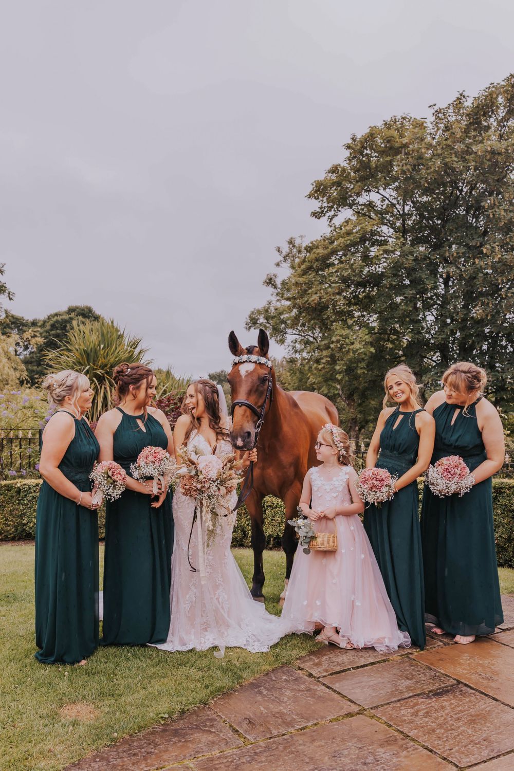 Wedding party in navy blue dresses poses with bride in outdoor garden setting with horse and pink floral bouquets.