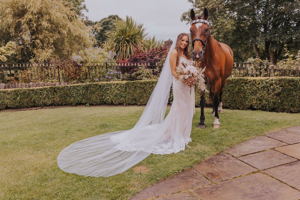 A horse stands next to someone in a flowing white wedding dress in a garden setting during a photo session.