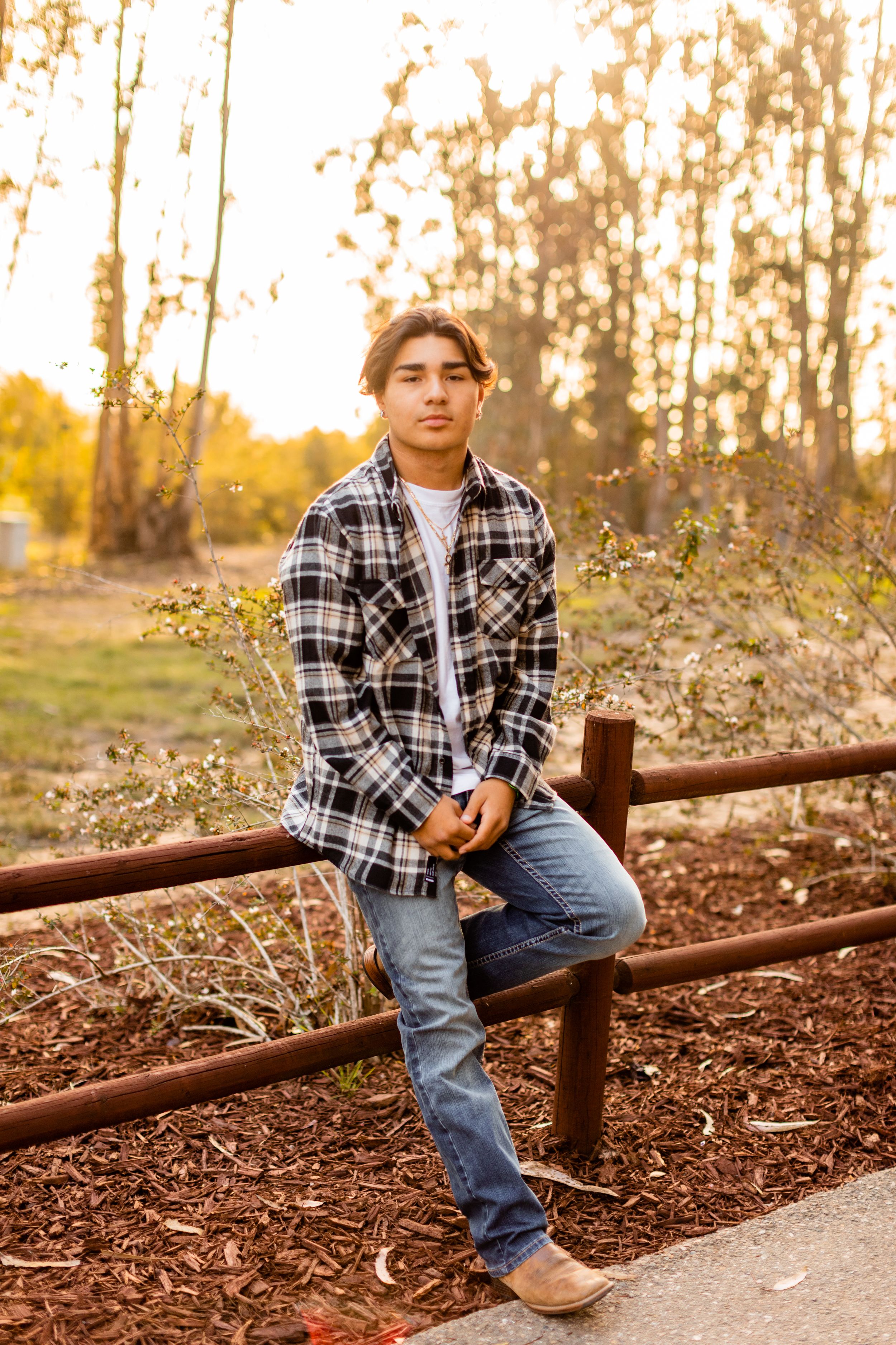A person in a plaid flannel shirt and jeans poses casually while leaning against a wooden fence in autumn.