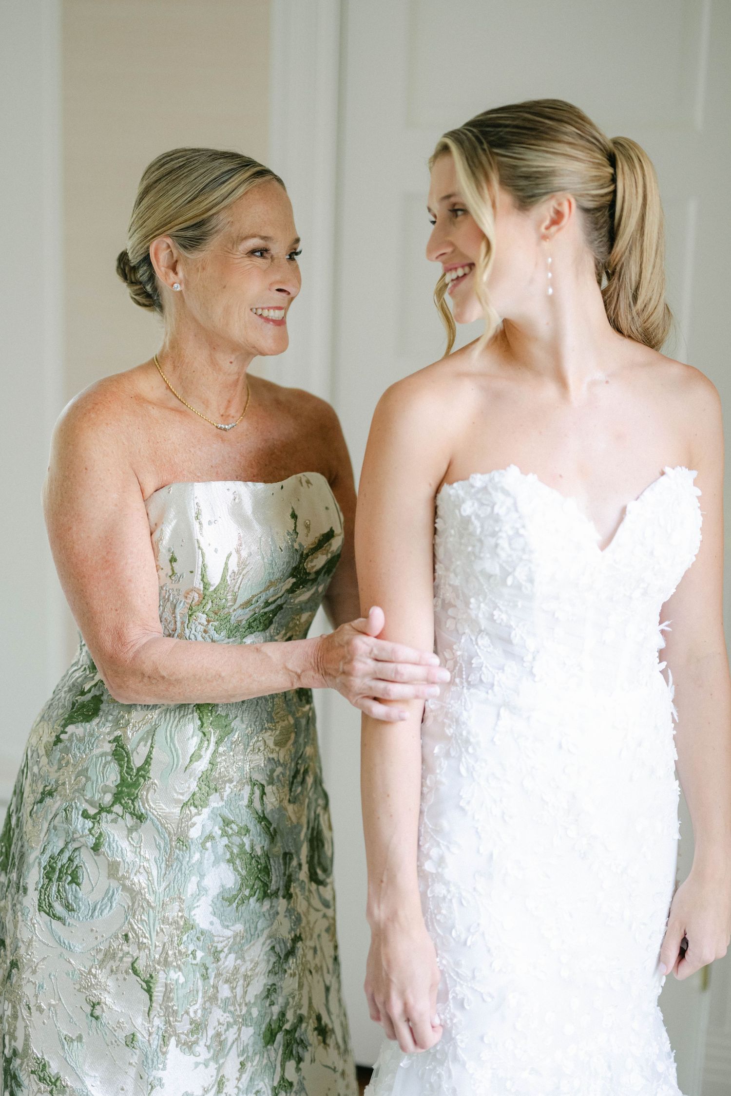 Two women share a joyful moment in a sun-lit room, one wearing a sage green pleated gown and one in a lace wedding dress. - Lake Forest