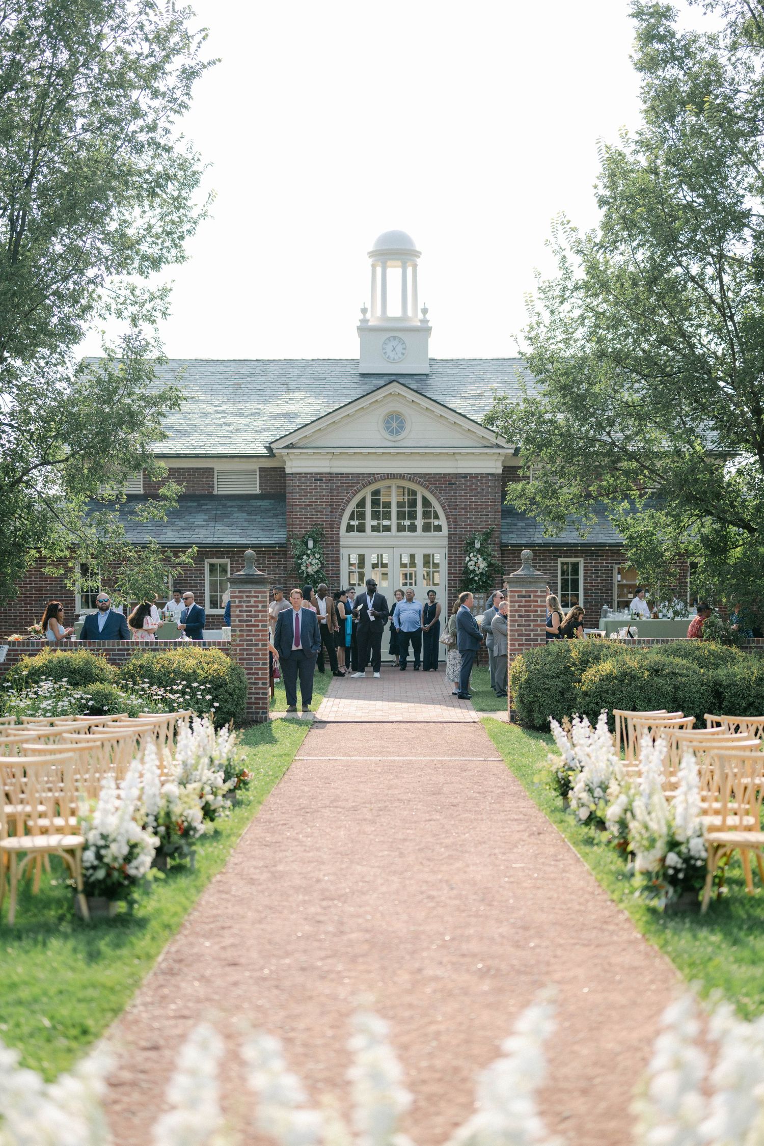 A rustic outdoor wedding venue with wooden chairs lining a gravel path leading to a colonial-style brick building. - Elawa Farm