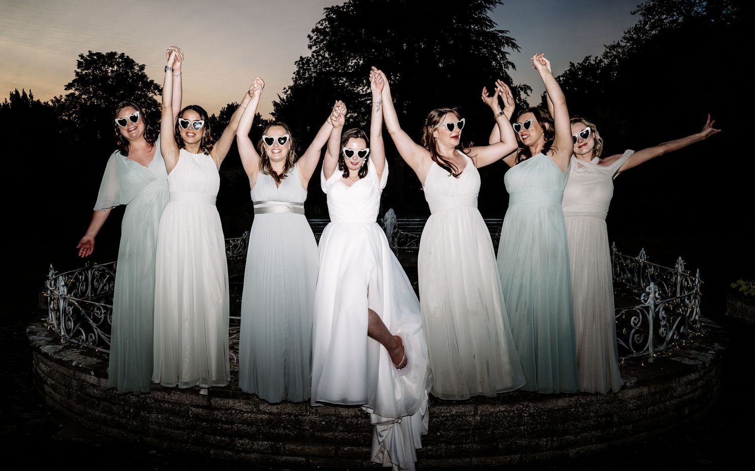 Silhouetted bridesmaids in light dresses posing with raised arms at dusk.