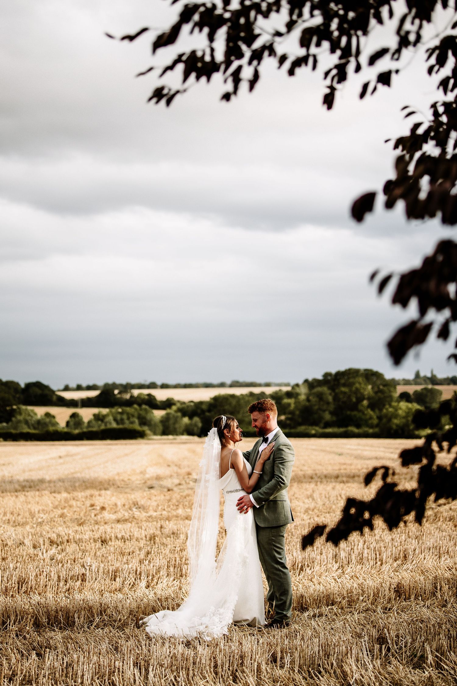 A couple shares a romantic moment in a golden wheat field under cloudy skies during their countryside wedding photoshoot.