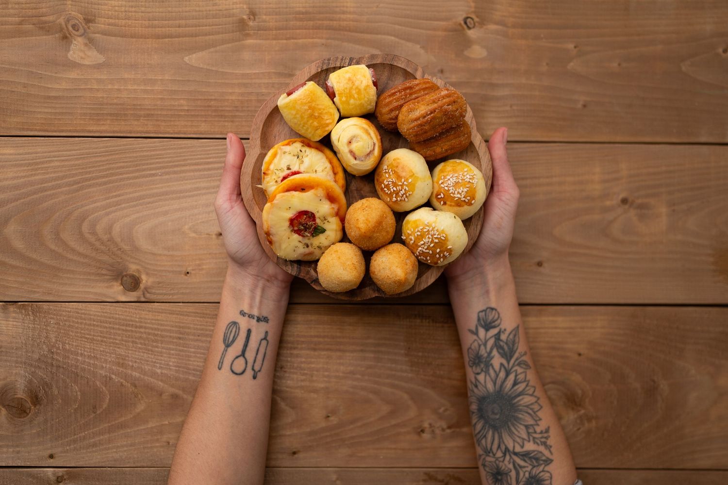 Tattooed hands holding a wooden bowl filled with freshly baked bread rolls and pastries on a rustic wooden surface.