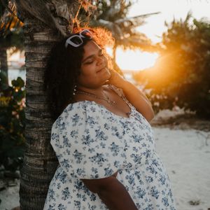 Woman in floral dress leaning against palm tree at golden hour sunset on tropical beach.