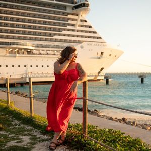 Woman in red dress poses by railing with MSC Seascape cruise ship docked behind her at sunset.