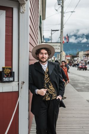 A young man in a vintage black suit, gold vest, and wide-brim hat leans against a building on a small-town street.
