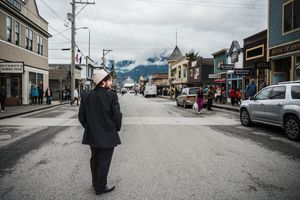 A person in a dark coat and white hat walks down a small town main street with mountains and cloudy skies in the background.