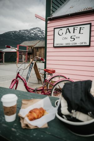 Red bicycle parked outside Cafe on 5th pink building with croissant and coffee on green table, snowy mountains backdrop.