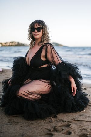 Woman in black tulle gown and sunglasses sitting on sandy beach with ocean waves in background.