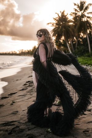 Woman in dramatic black feathered gown poses on tropical beach at sunset wearing sunglasses with palm trees behind her.