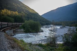 A scenic railway bridge crosses a rushing mountain river surrounded by autumn forests and misty peaks at dusk.