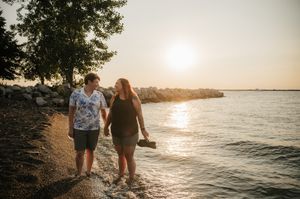 Couple holding hands walking along a rocky shoreline at golden sunset with calm water in the background.