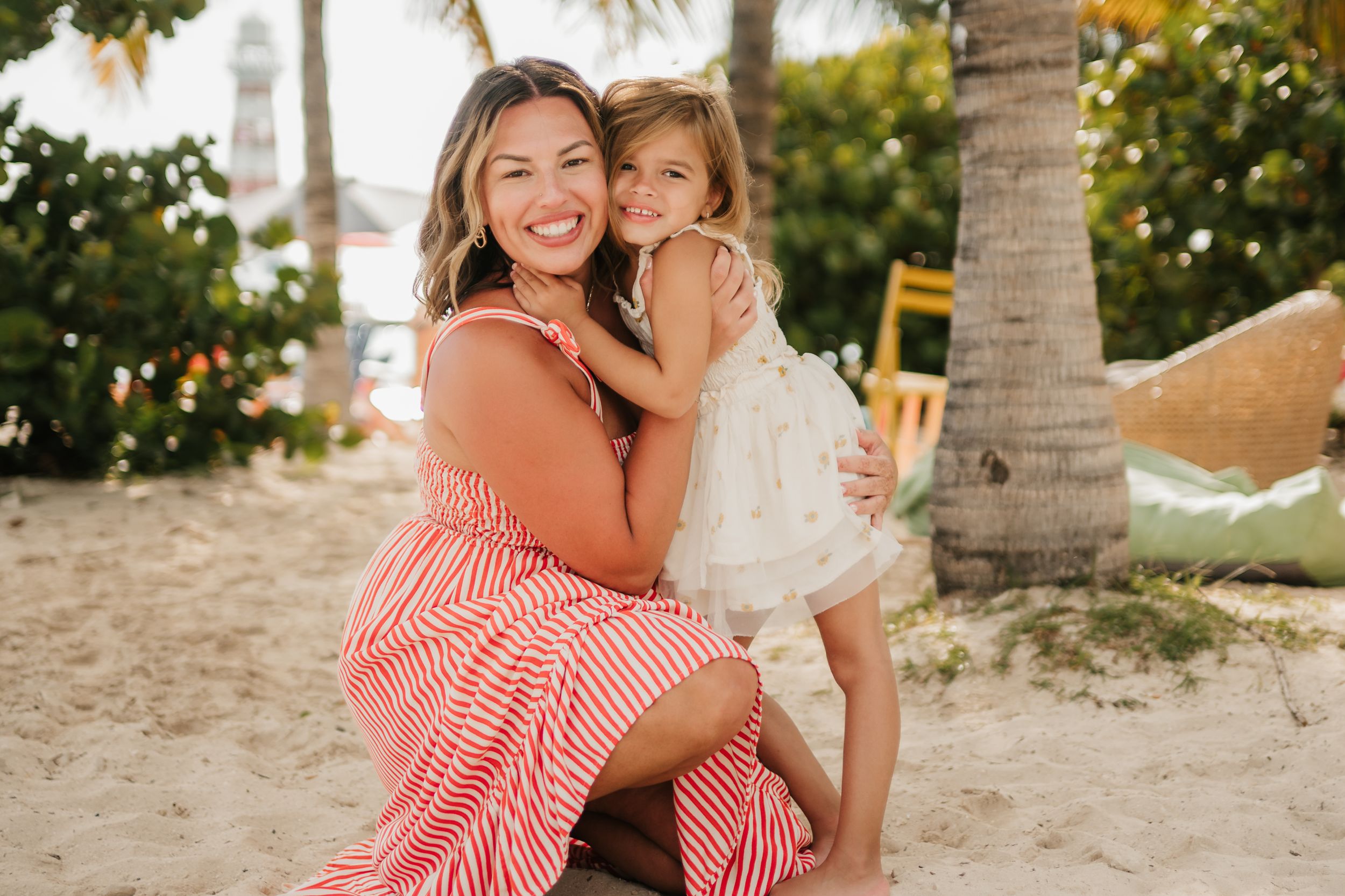 A smiling woman in a red striped dress hugs a young girl in white on a sandy beach near a palm tree.