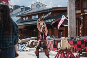 Lumberjack performer raises chainsaw overhead at outdoor timber sports show near rustic log cabin venue.