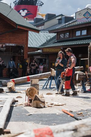 Lumberjack show performers with chainsaw near log carvings, concessions stand and Disney cruise ship visible in background.