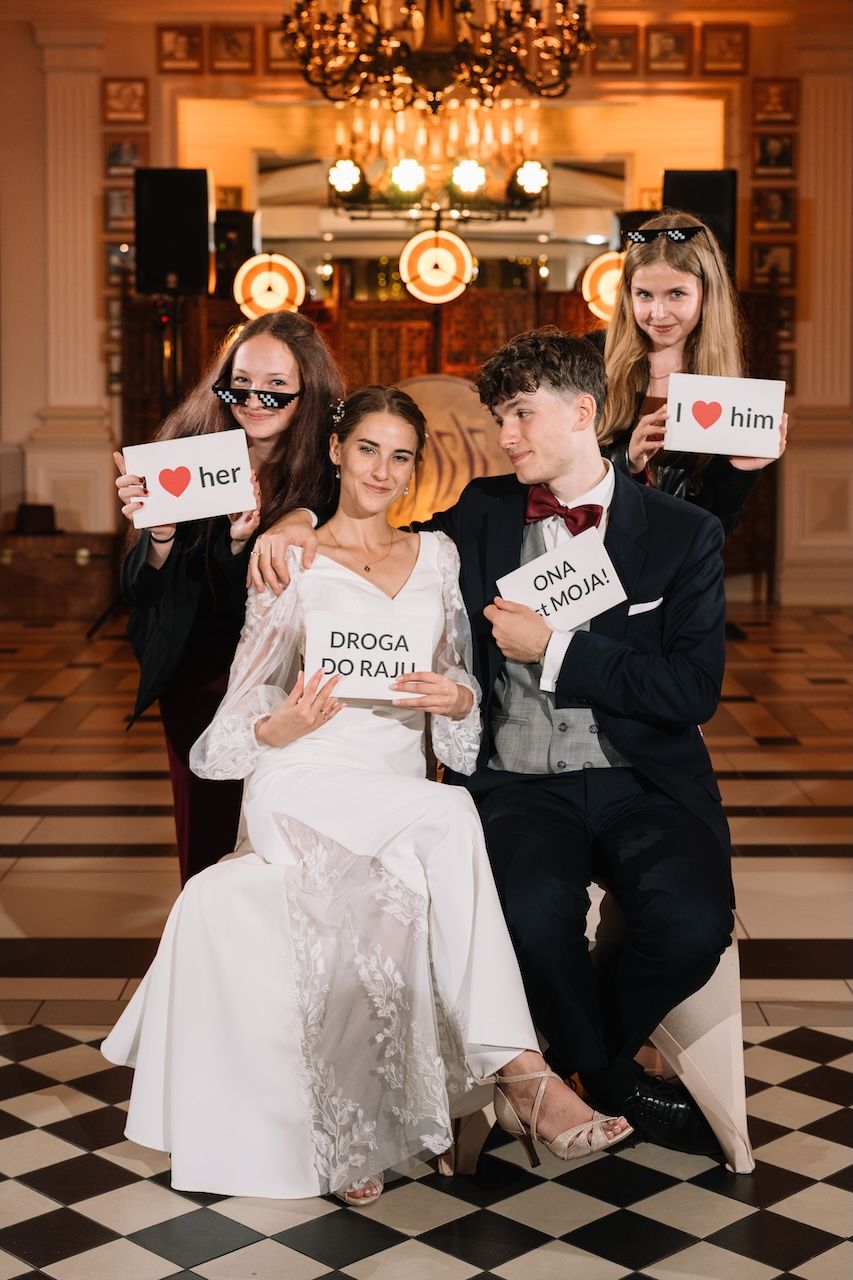 Wedding party posing with fun signs including I love her, I love him, and Droga Do Raju at elegant reception.