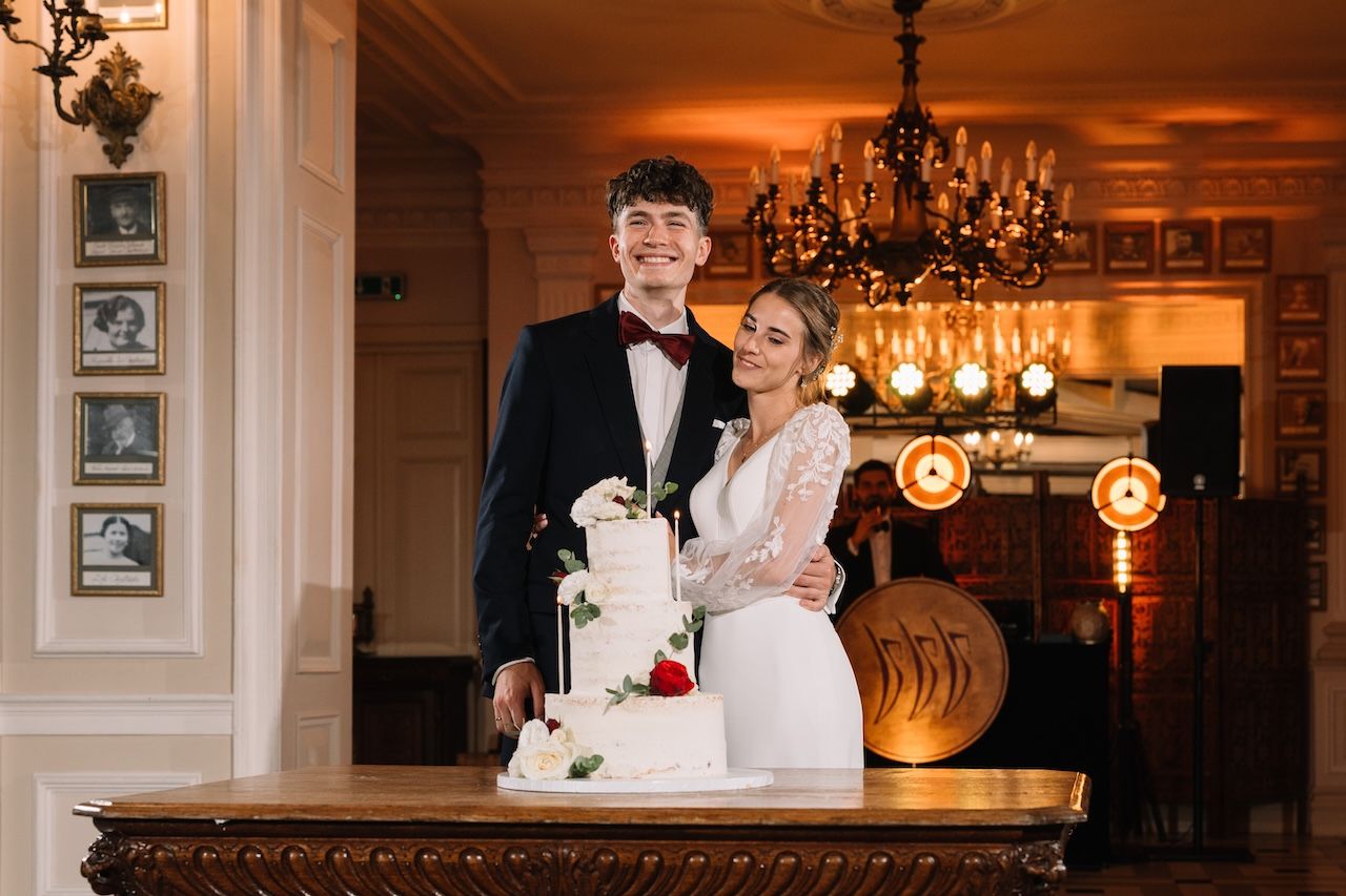 Newlyweds smiling together beside a white tiered wedding cake in a warmly lit elegant venue.