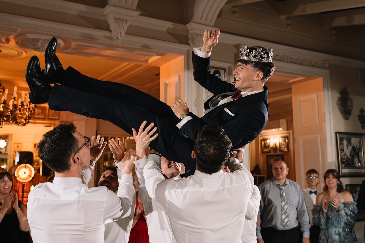 Groomsmen tossing the groom into the air wearing a crown during a lively wedding celebration.