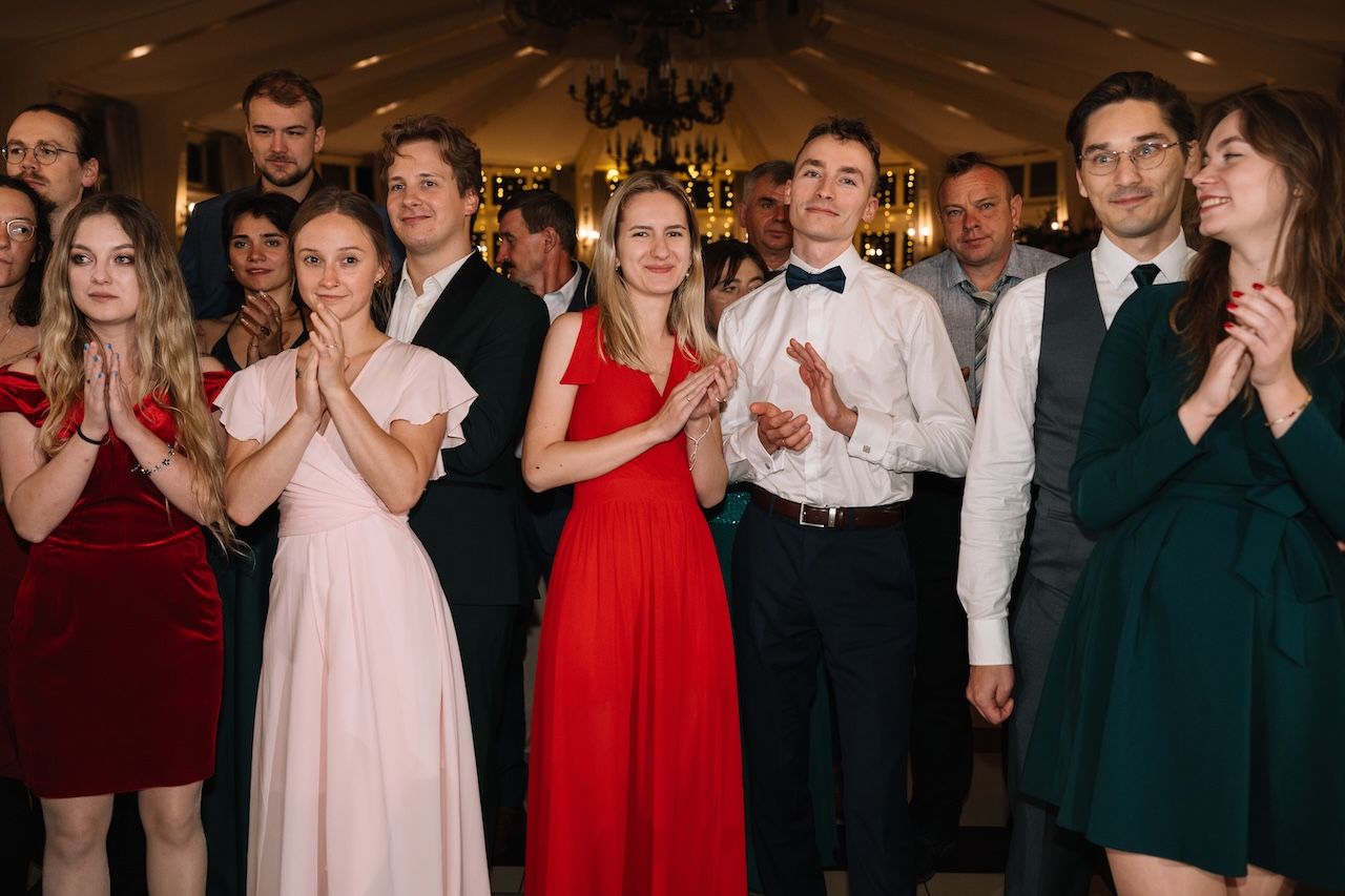 Elegantly dressed wedding guests clap and smile during indoor reception celebration.