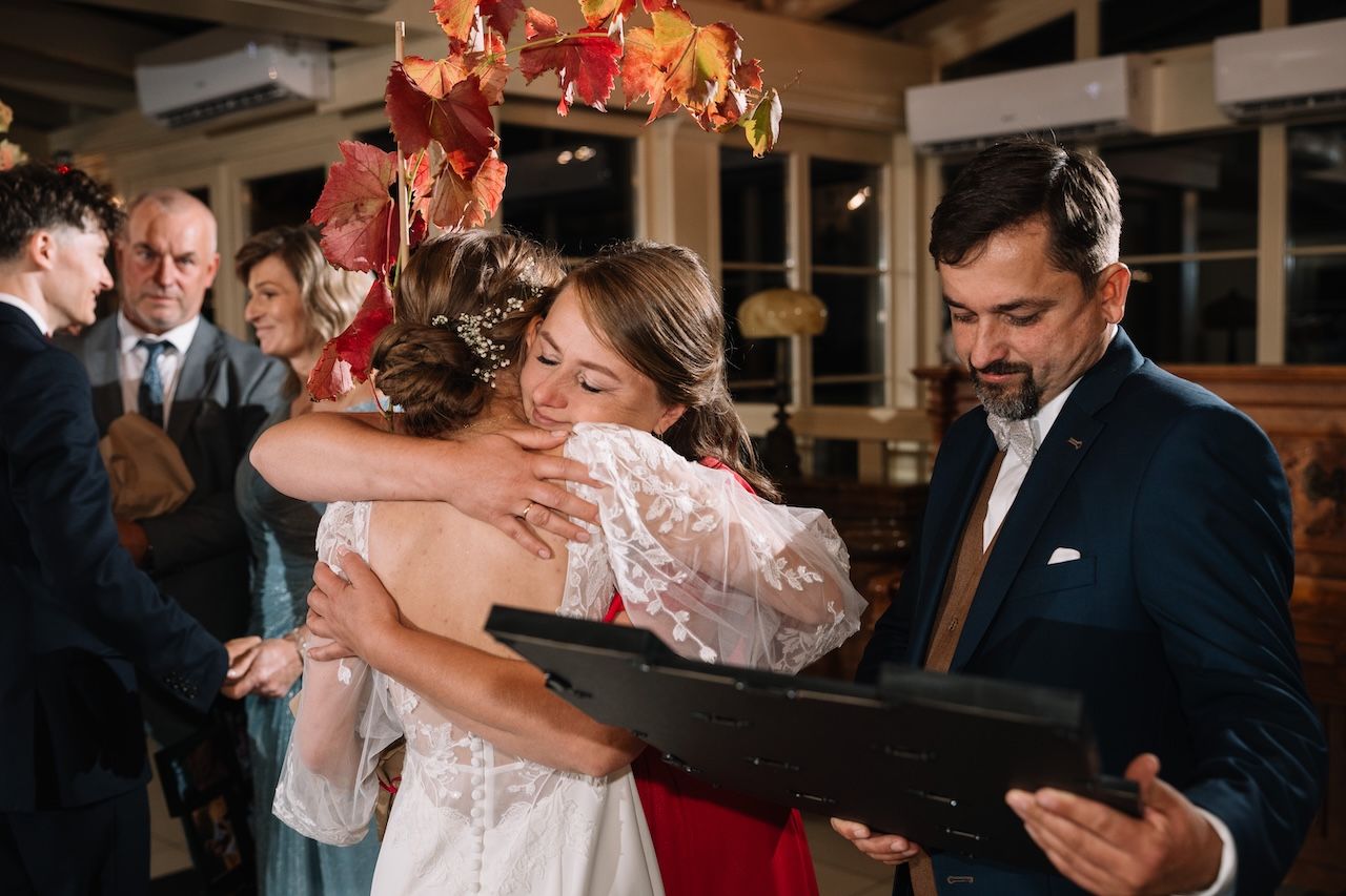 Bride in white lace gown shares emotional hug with guest at wedding reception.