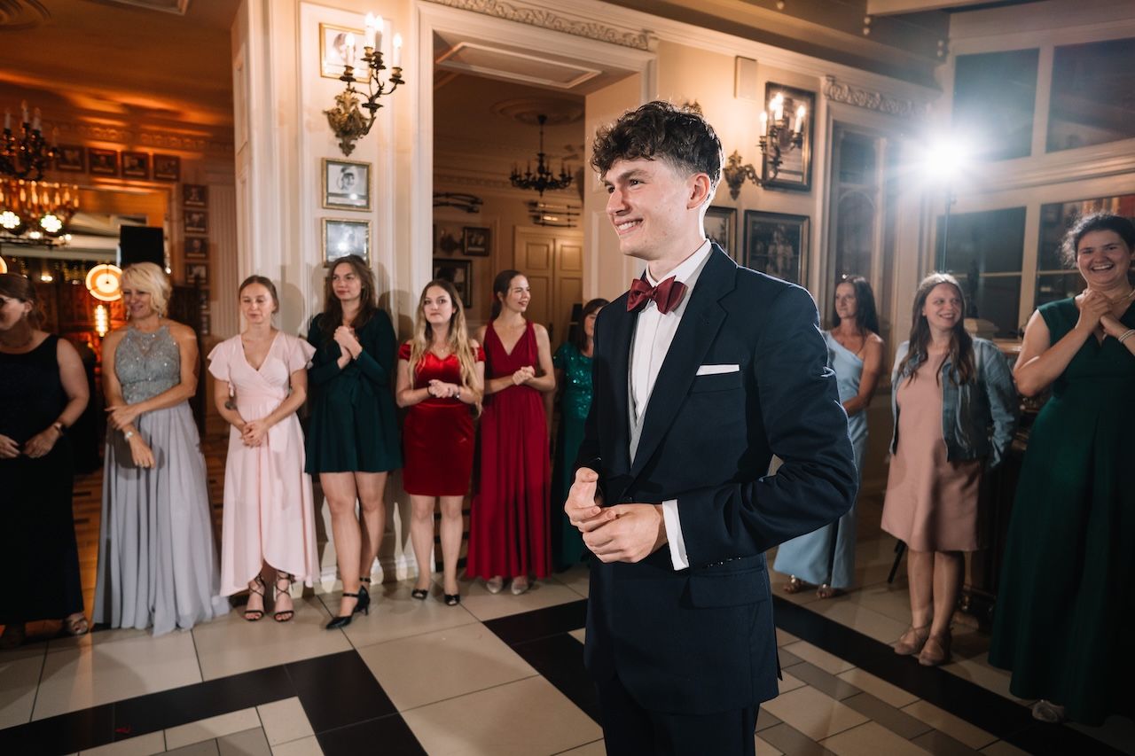 Groom in navy tuxedo smiles on dance floor surrounded by wedding guests.