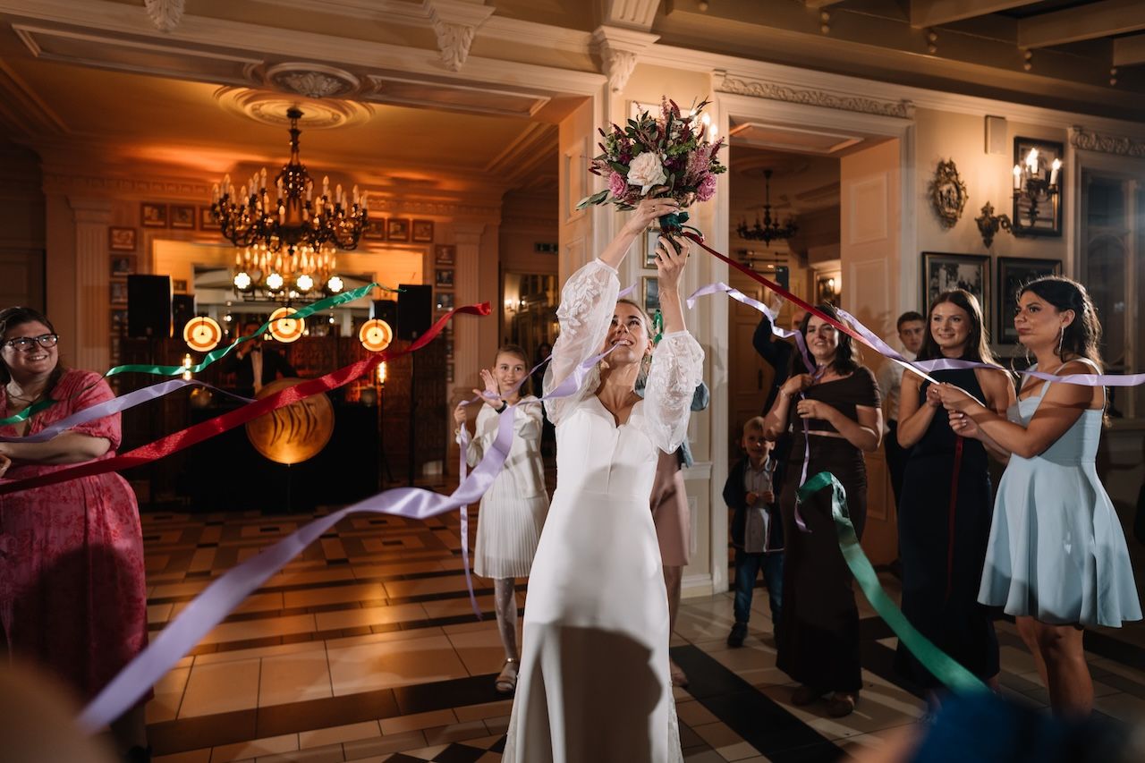 Bride in white gown tosses bouquet to guests with ribbons at wedding reception.