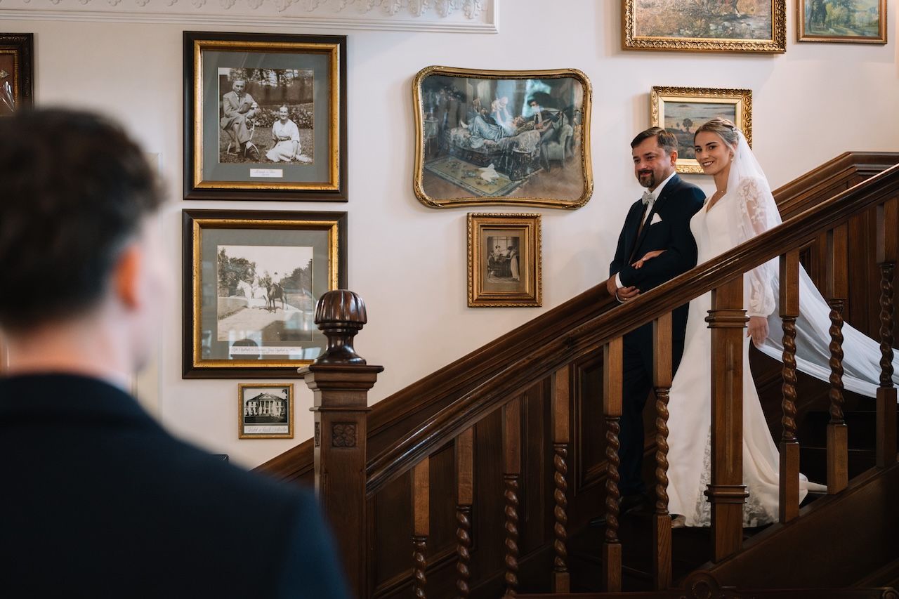 Elegant bride and groom descending ornate wooden staircase adorned with framed artwork at wedding.
