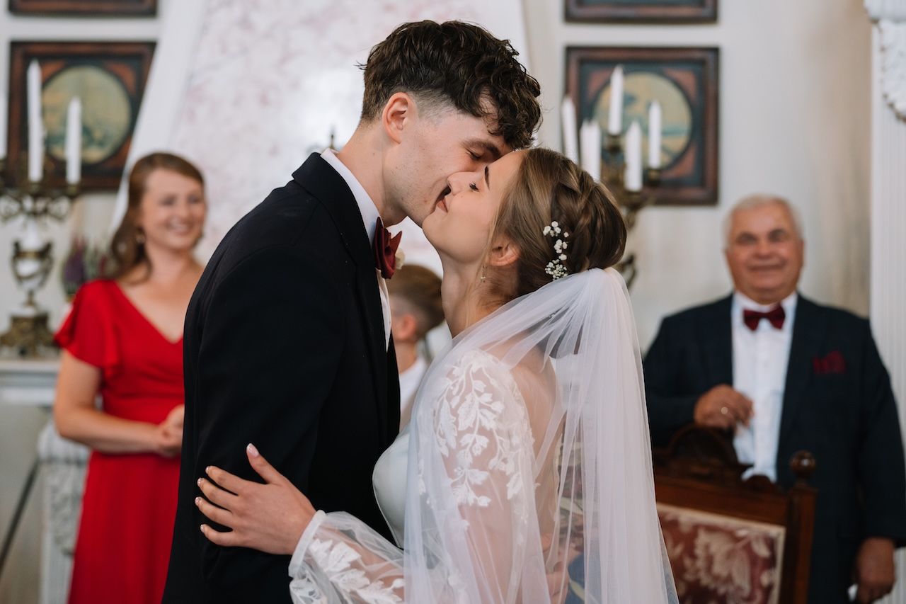 Bride and groom sharing romantic kiss during wedding ceremony with smiling guests in background.