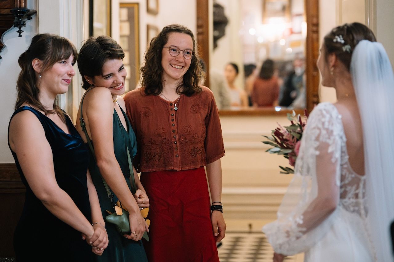 Bride in lace veil greeting smiling bridesmaids in elegant dresses at wedding reception venue.
