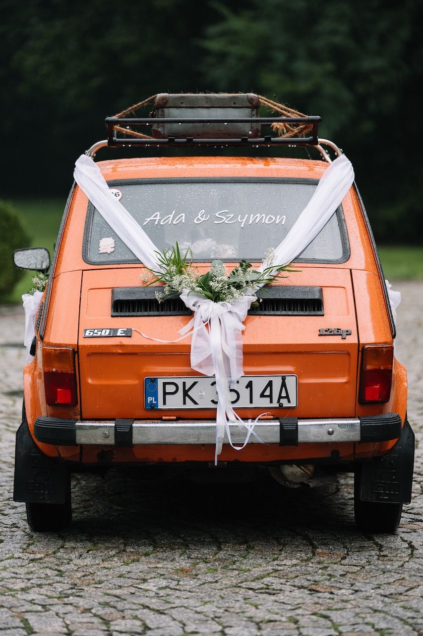 Orange vintage wedding car decorated with white ribbons and flowers, license plate PK 3514A, with Ada & Szymon written on rear window.