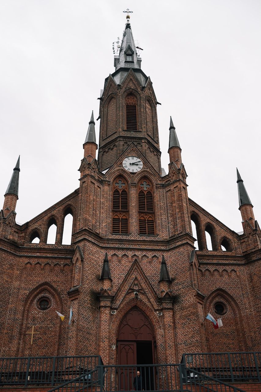 Gothic red brick church with tall clock tower, pointed spires, and arched entrance doorway against overcast sky.