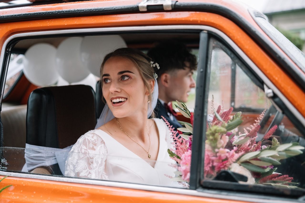 Smiling bride in white lace dress sitting in an orange vintage car with groom visible in background.