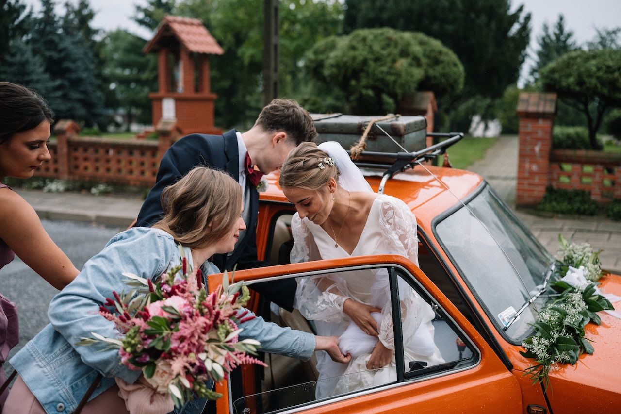 Wedding party helping bride in white lace dress exit an orange vintage car outside a church.