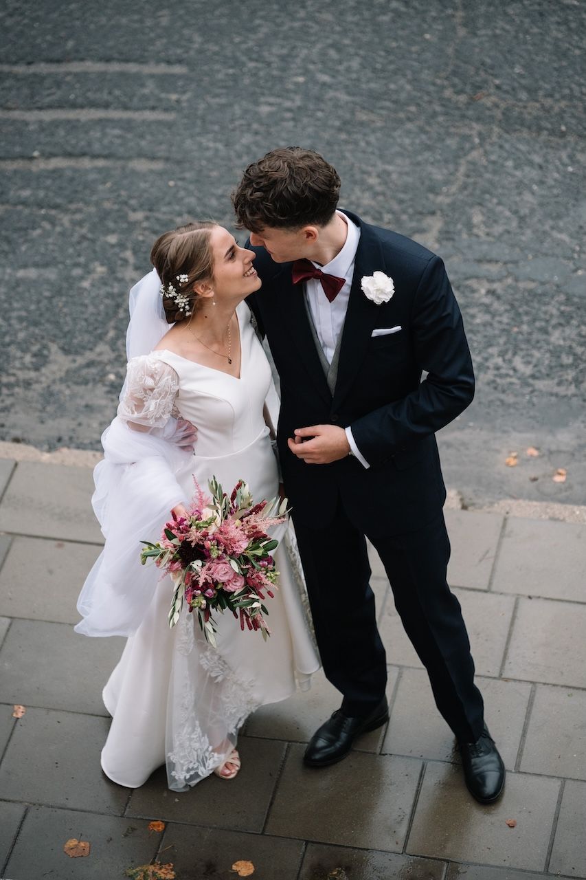 Bride and groom sharing a romantic moment on a rainy sidewalk, bride holding a pink and red floral bouquet.
