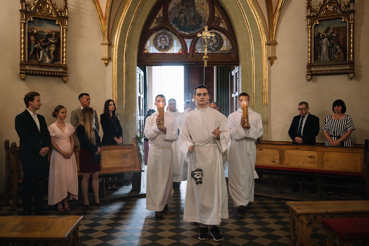 Wedding ceremony processional with altar servers in white robes leading guests through an ornate church entrance.