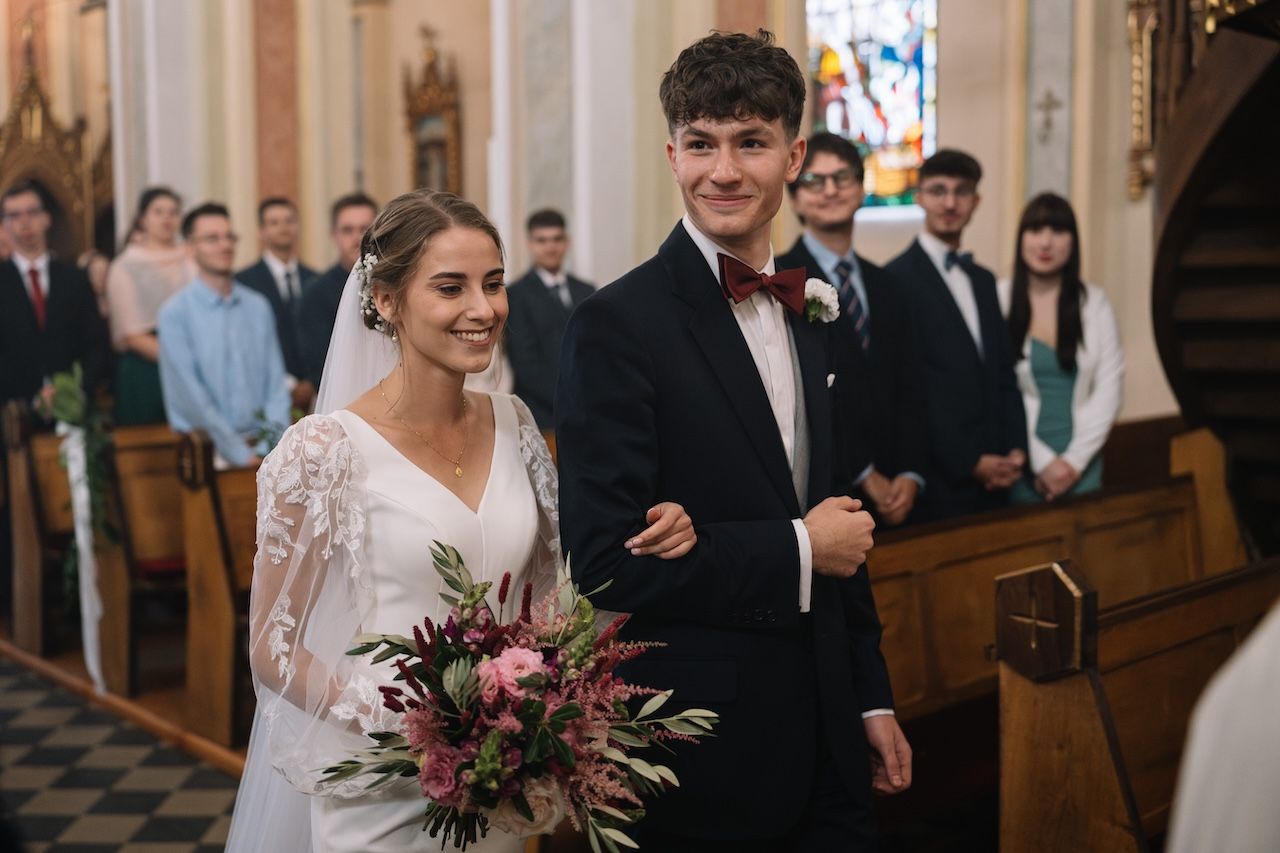 Smiling bride and groom walking down the church aisle together, bride holding a pink and red bouquet.
