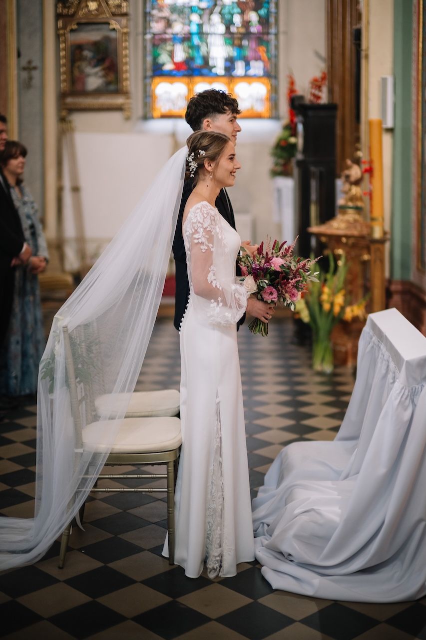 Bride in elegant white lace gown with long veil holds bouquet during church wedding ceremony.