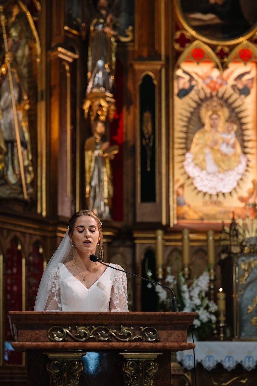 Beautiful bride in white lace dress speaks at church podium during wedding ceremony.