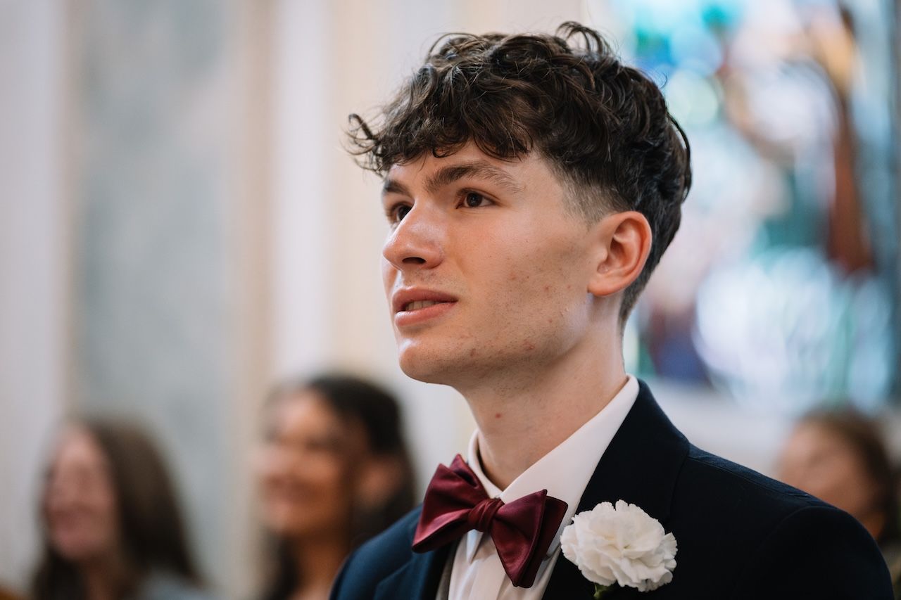 Young groom in navy suit with burgundy bow tie watches wedding ceremony emotionally.