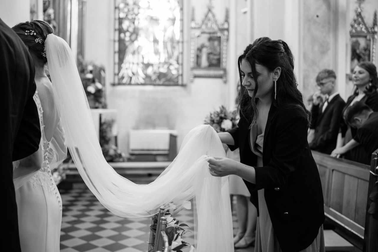 Black and white photo of bridesmaid arranging bride's long veil in church aisle.