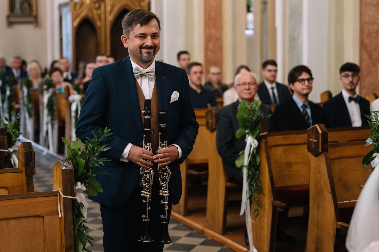 Smiling musician holds clarinet while standing in flower-decorated church aisle at wedding.