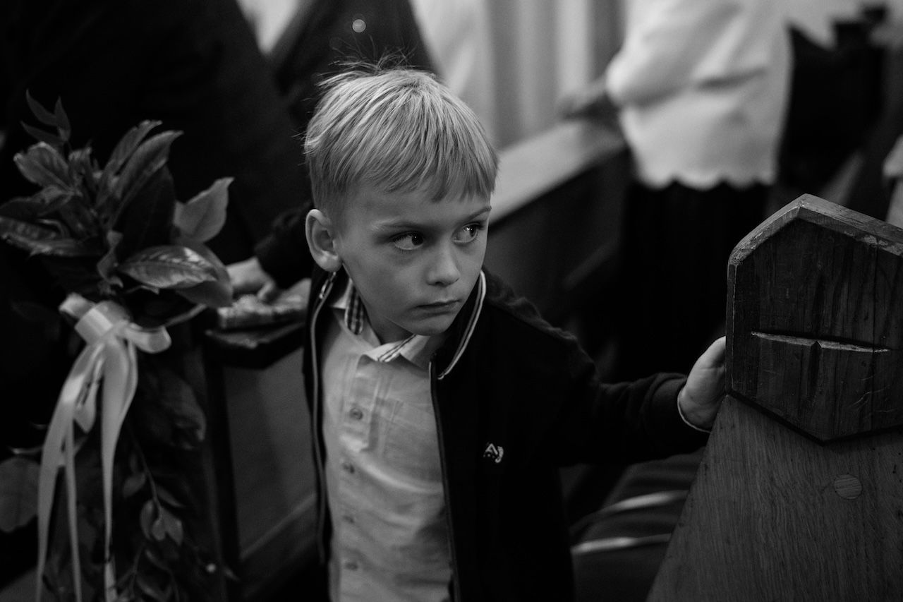 Serious young blond boy in suit stands by church pew during wedding ceremony.